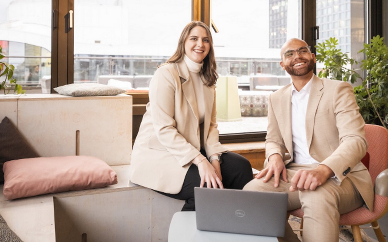 Zwei Personen in heller Businesskleidung sitzen in einem modernen Raum mit Laptop, großen Fenstern, Pflanzen und Blick auf die Stadt.