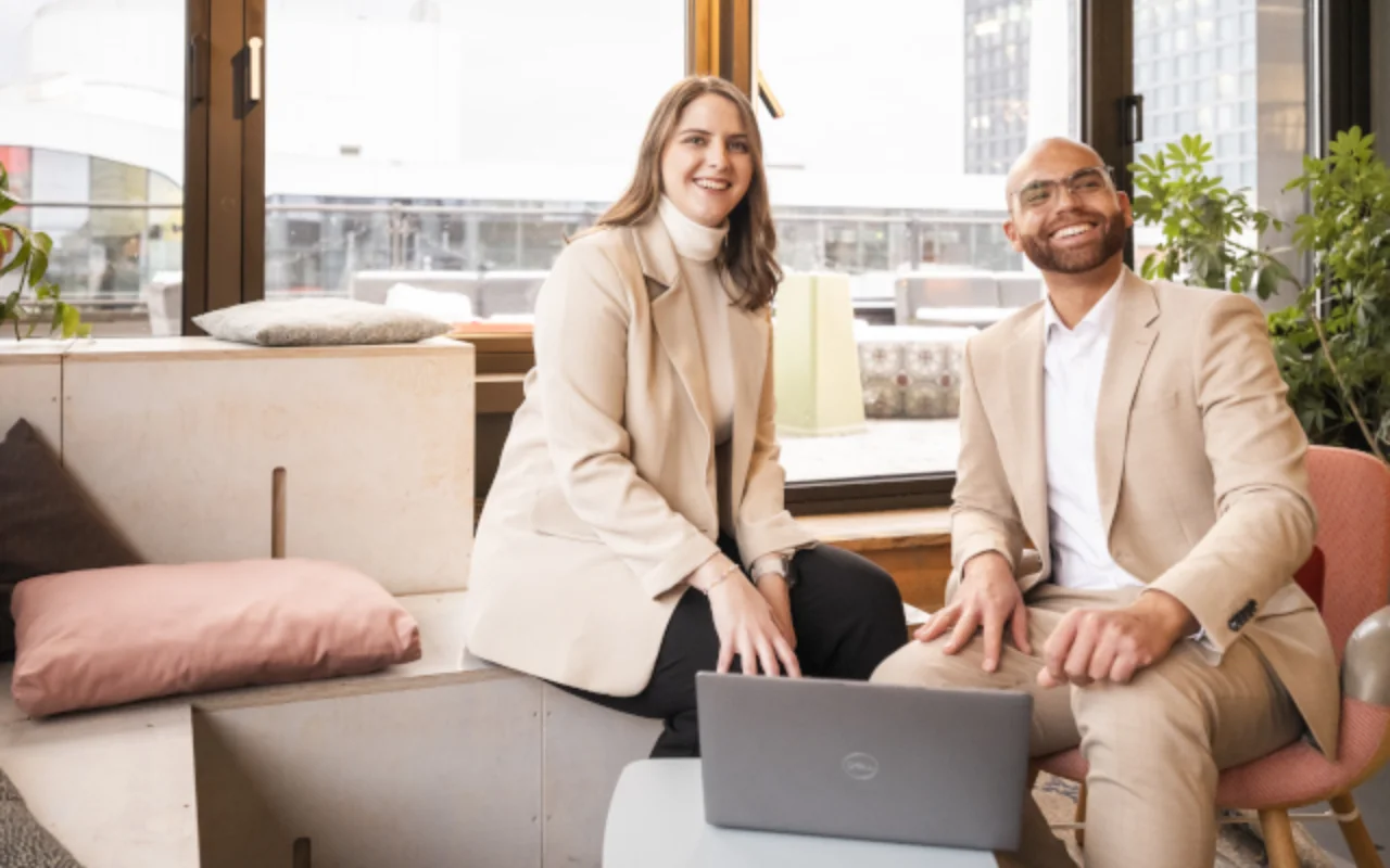 Zwei Personen in heller Businesskleidung sitzen in einem modernen Raum mit Laptop, großen Fenstern, Pflanzen und Blick auf die Stadt.