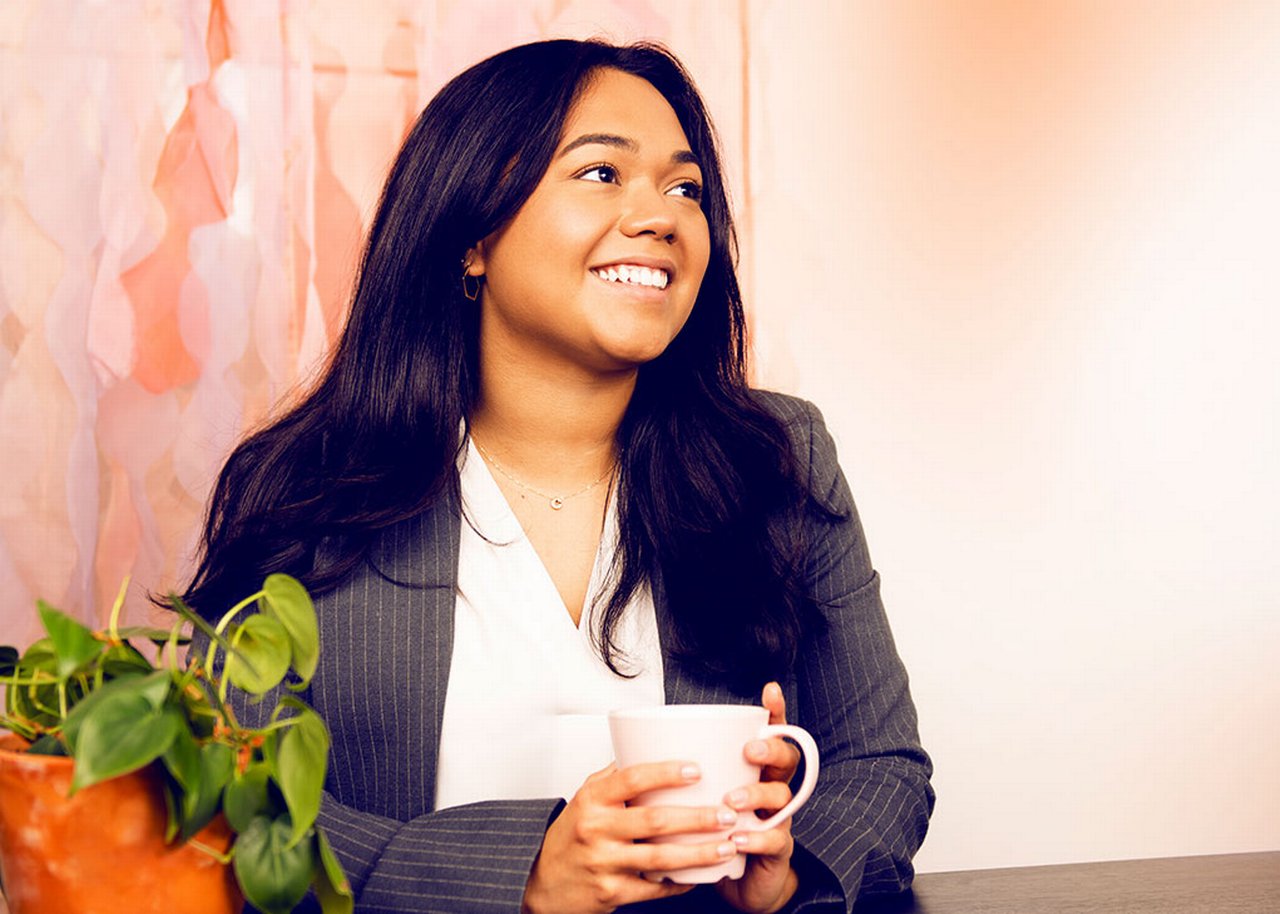 Camila lächelt mit einer Tasse in der Hand, neben ihr eine Topfpflanze
