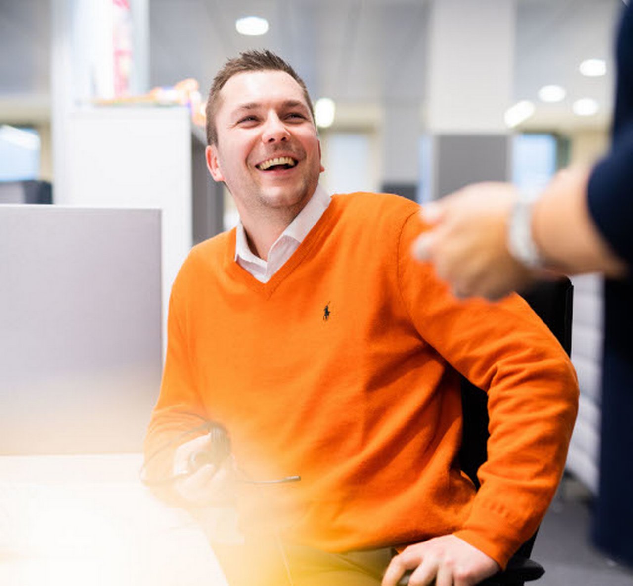 Andreas unterhält sich lachend im Büro mit einem Kollegen. Er trägt einen orangen Pullover.
