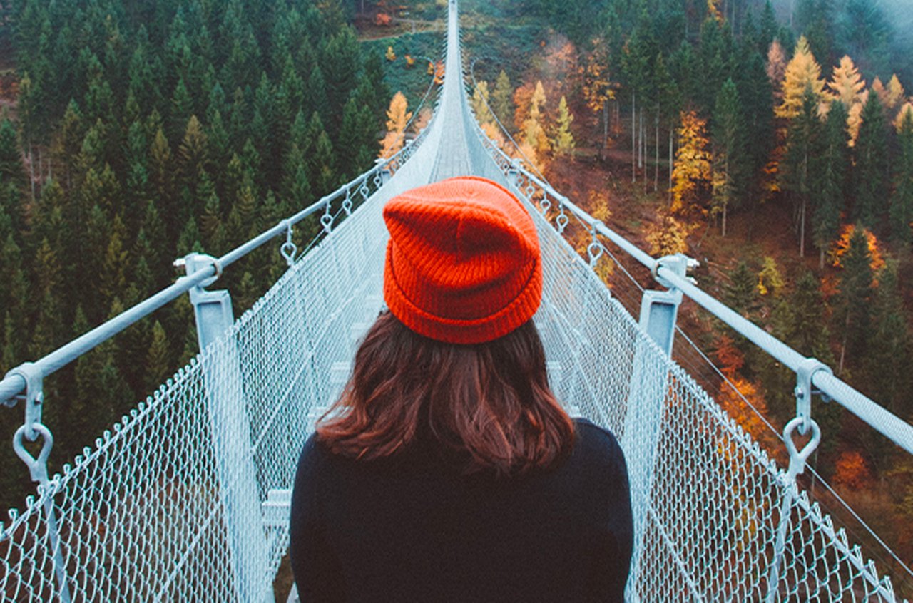 A woman with a red cap is walking on a bridge that leads over an autumn forest