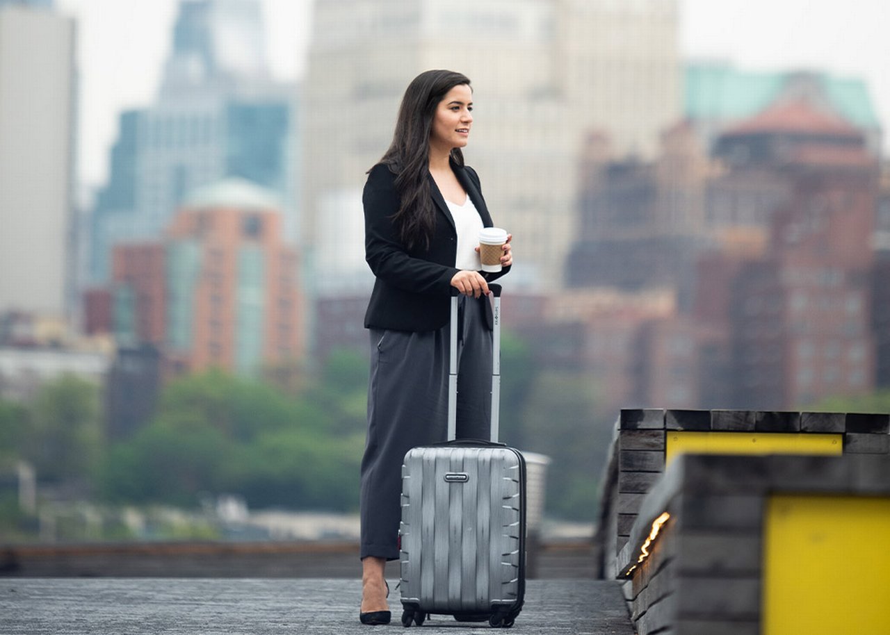 Monica is standing in front of skyscrapers with a suitcase and a coffee in her hand
