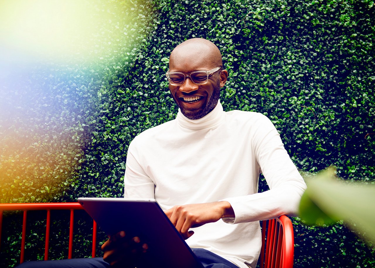 Kofi with tablet laughing on a red bench in front of a box hedge