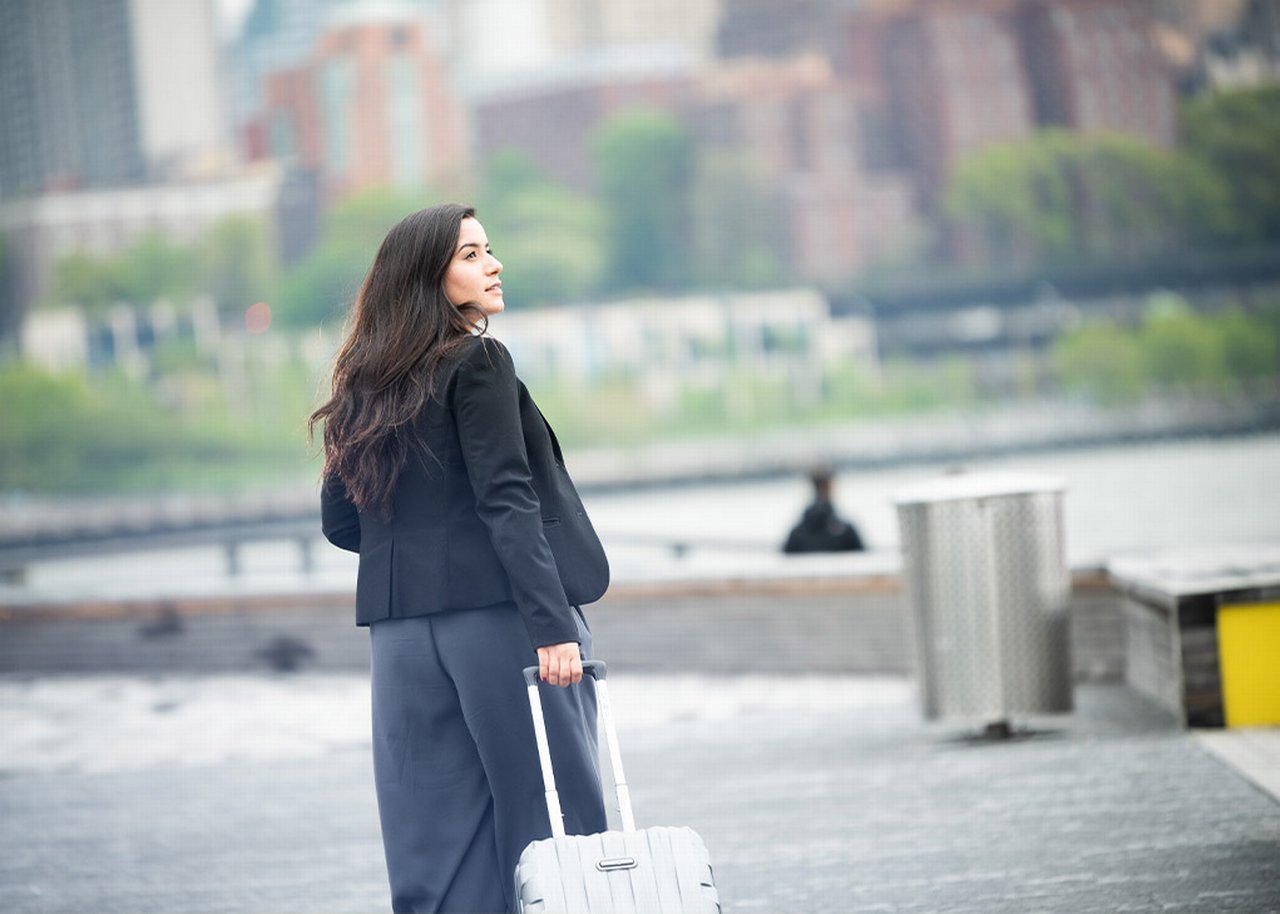 Monica is standing out in the open with a suitcase in her hand and her back partially turned