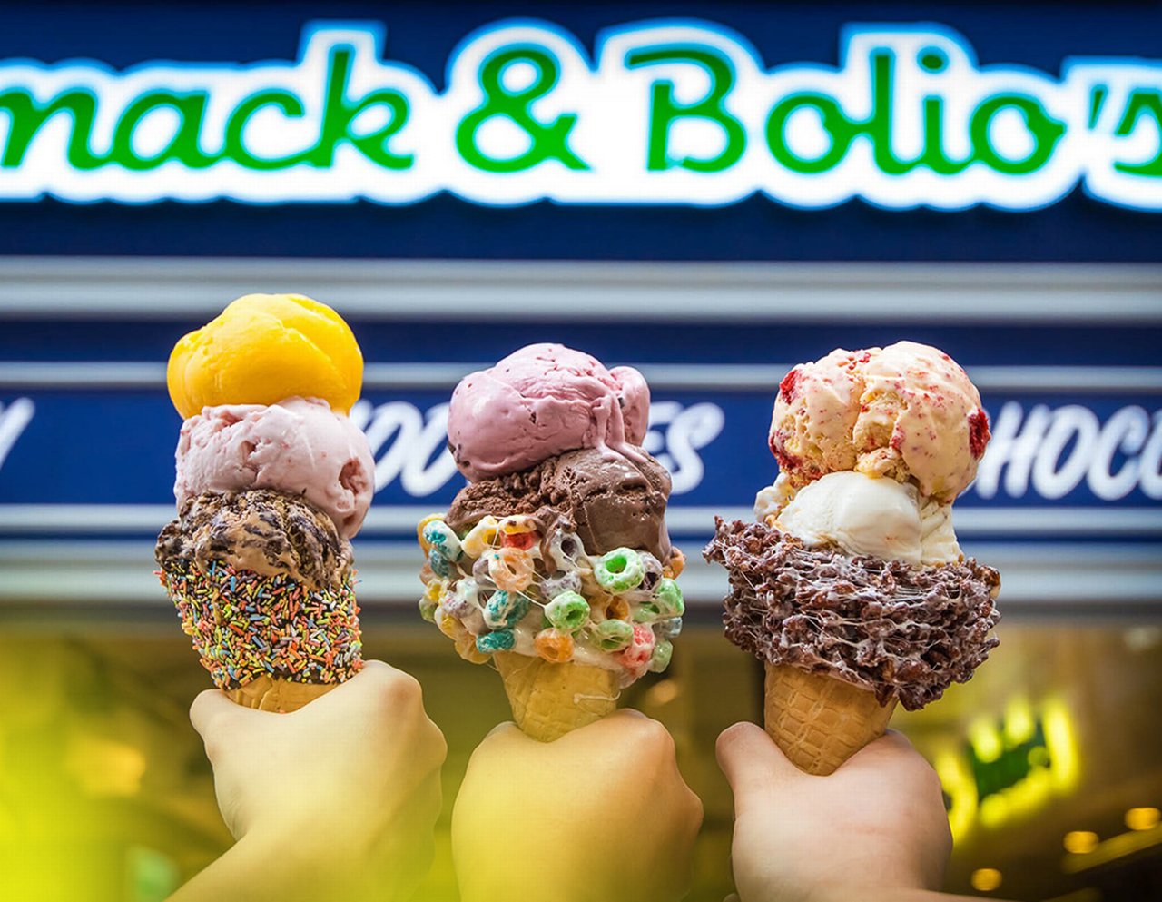 Three hands each holding Ice cream cones with different variety ice creams outside of an ice cream parlour