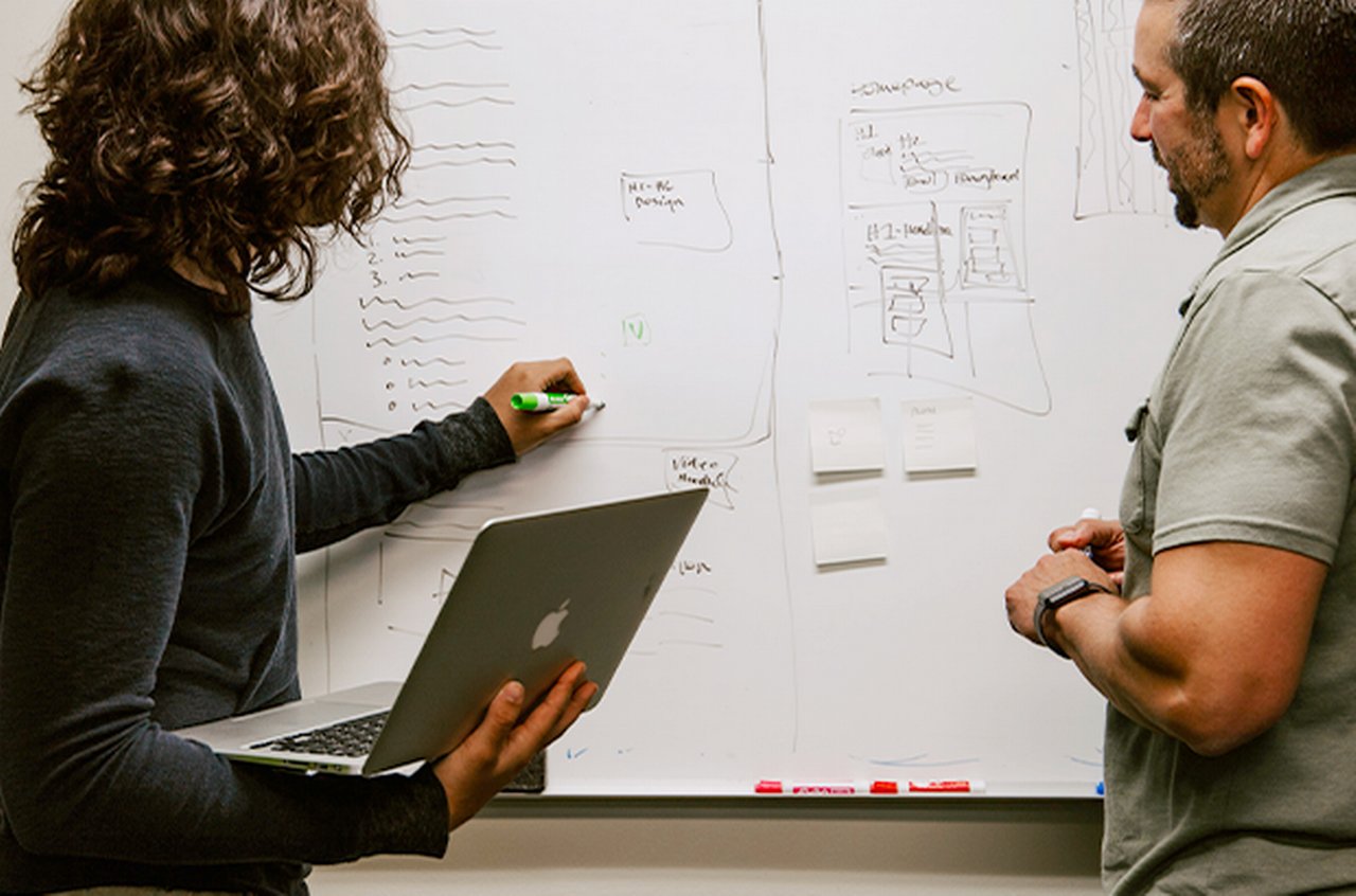 Two people at a whiteboard; a woman with a laptop in her arms writes something on it