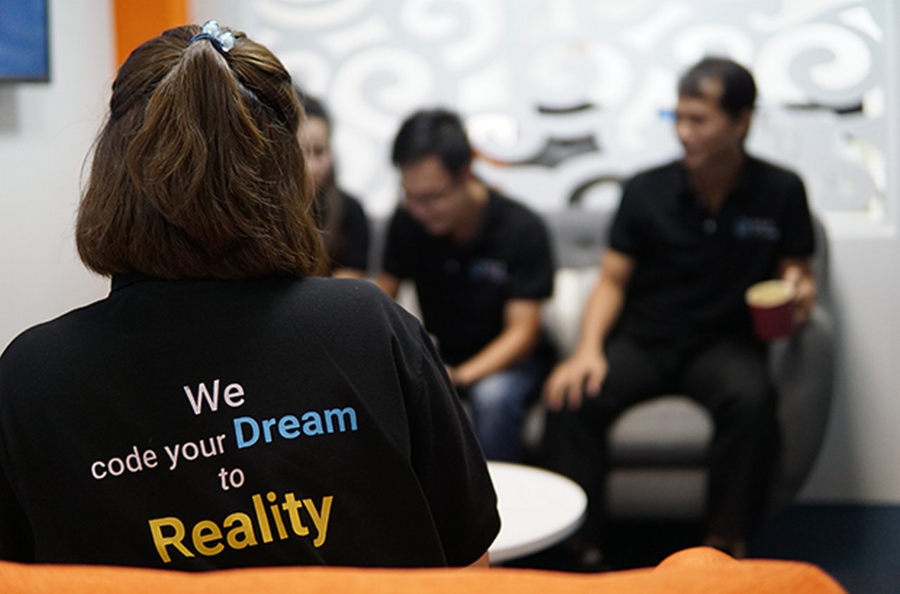 Woman in a meeting wearing a t-shirt that says ${esc.q}We code your Dream to Reality${esc.q}