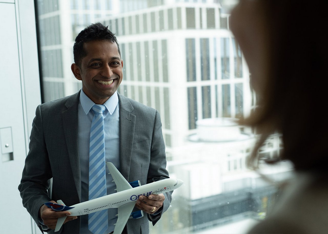 Janagran smiles and stands next to a window while holding a model airplane