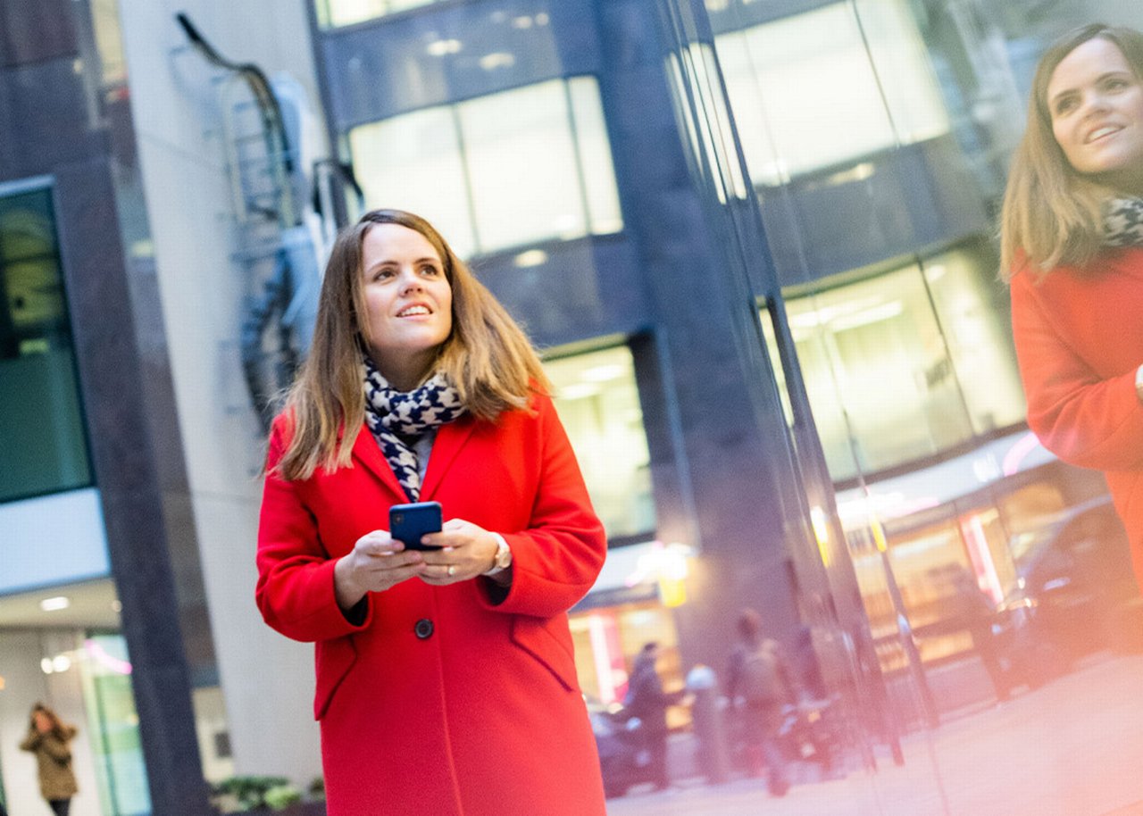 Sarah steht mit dem Handy in der Hand vor einem Glasgebäude