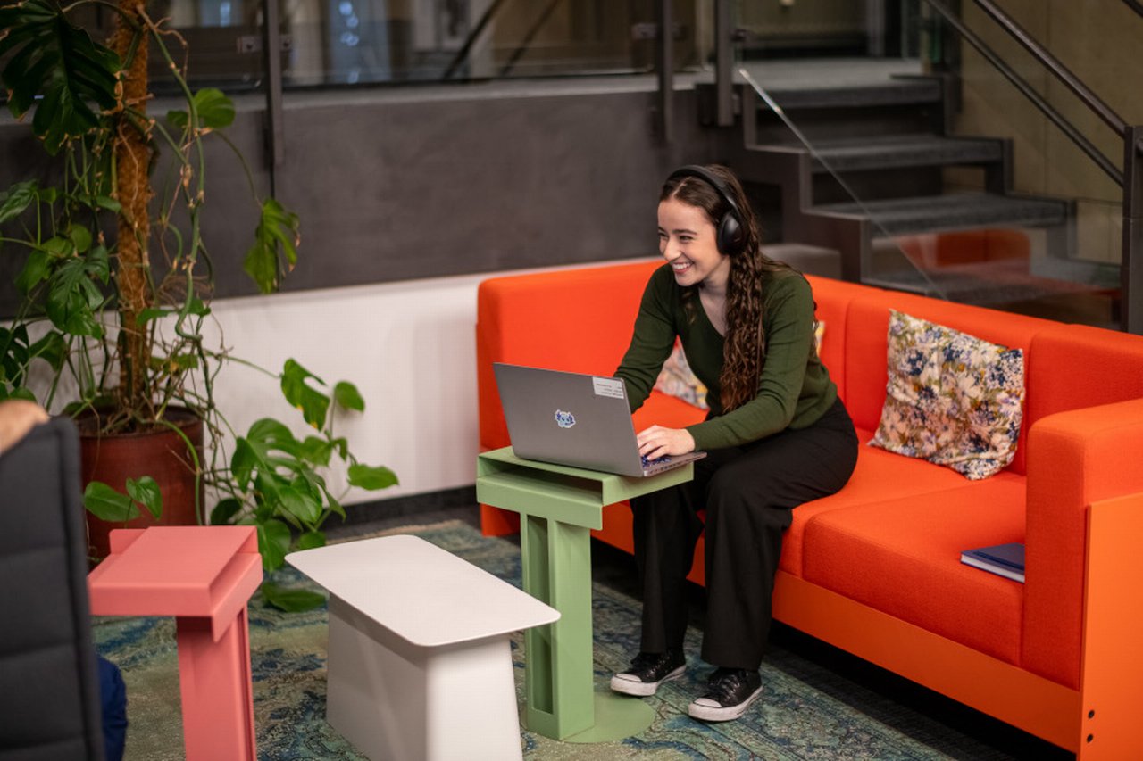 Person mit Kopfhörern arbeitet auf einem Laptop, sitzt auf einem orangefarbenen Sofa; im Hintergrund Treppe, Pflanzen und bunte Tische.