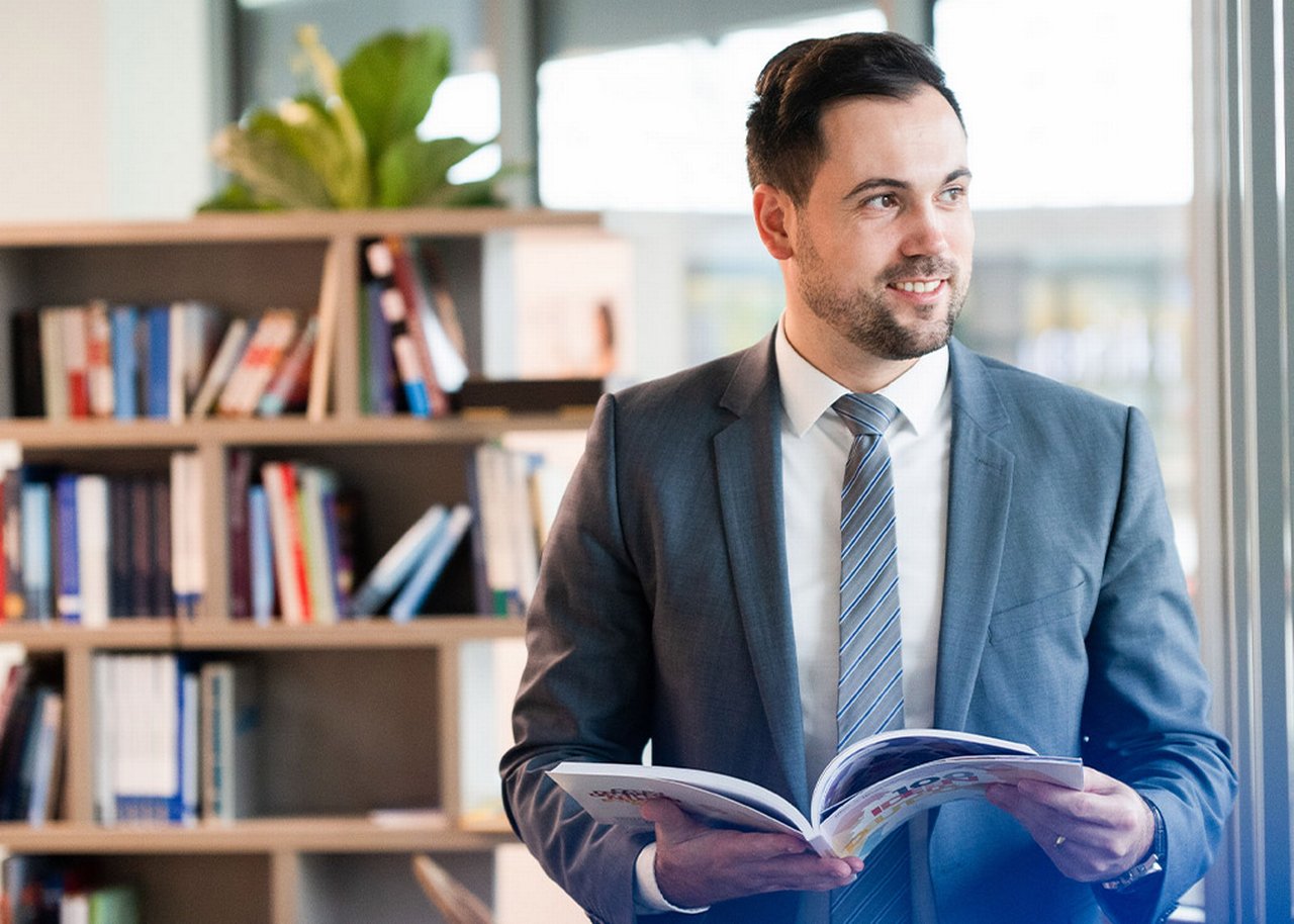 Semi steht mit einem Buch in der Hand vor einem Bücherregal