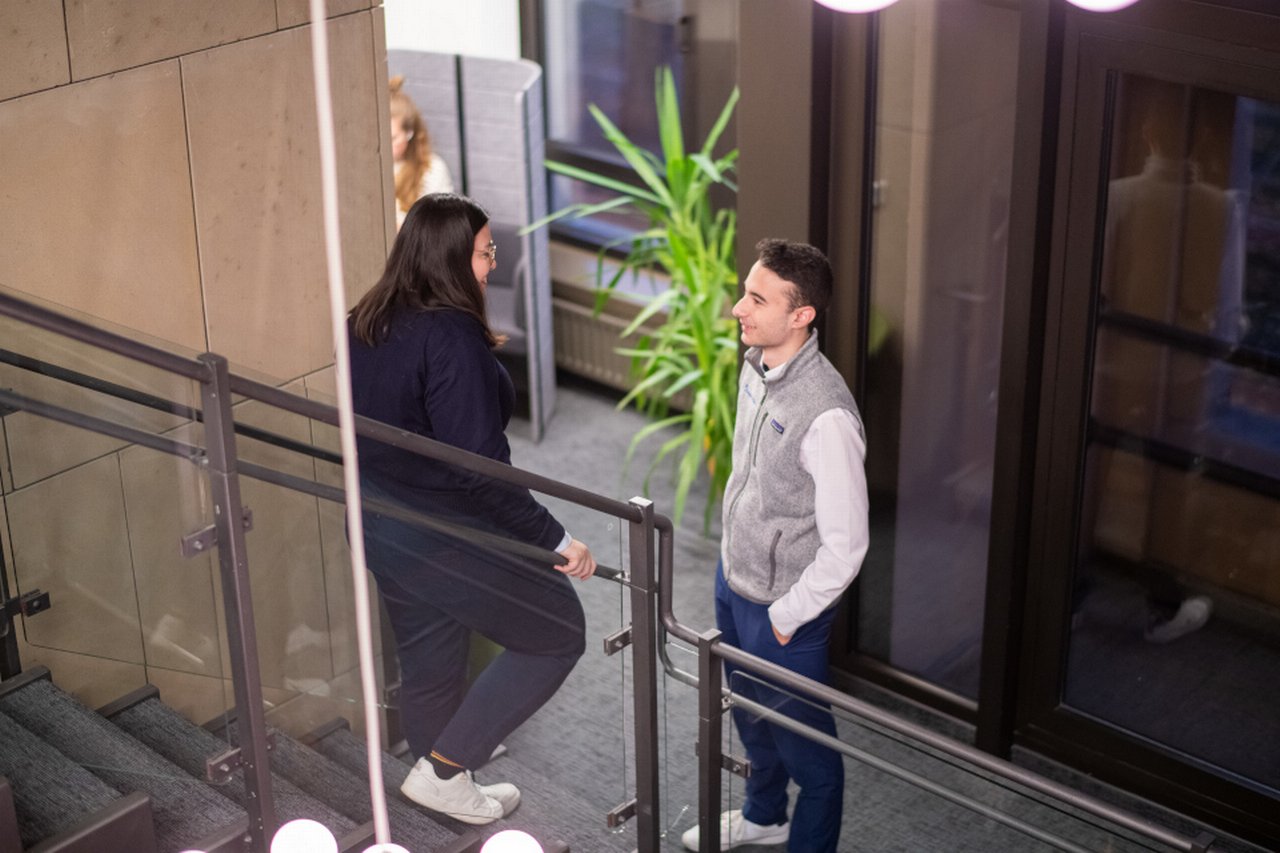 Zwei Personen unterhalten sich auf einer Treppe in einem Gebäude; im Hintergrund sind eine große Pflanze und Büromöbel zu sehen.