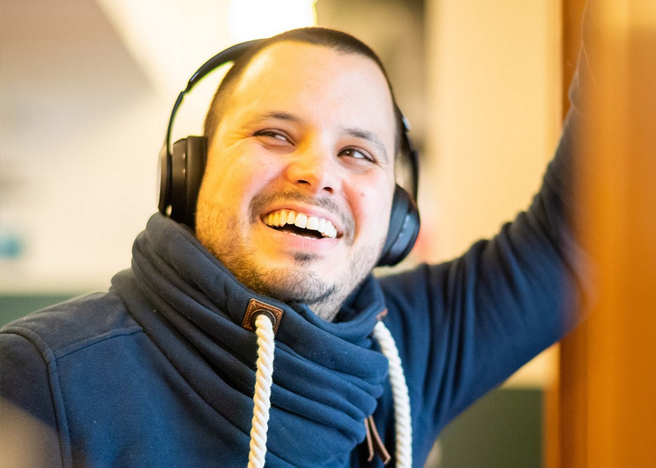 Ralf is leaning against a wall smiling with headphones on