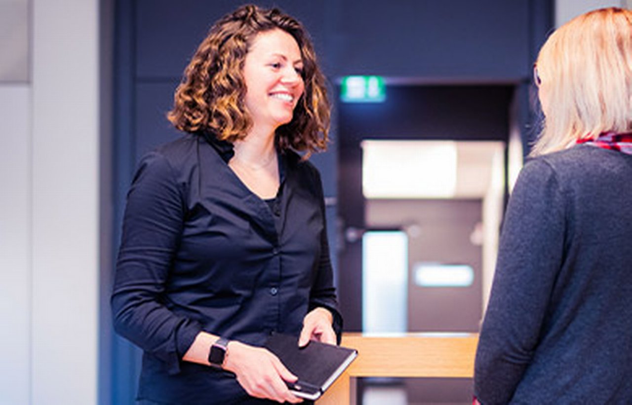 Erin has a notebook in her hand and is smiling with a colleague