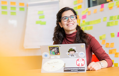 Sofie smiles and has opened her laptop, on which there are many stickers. A whiteboard with notes can be seen in the background